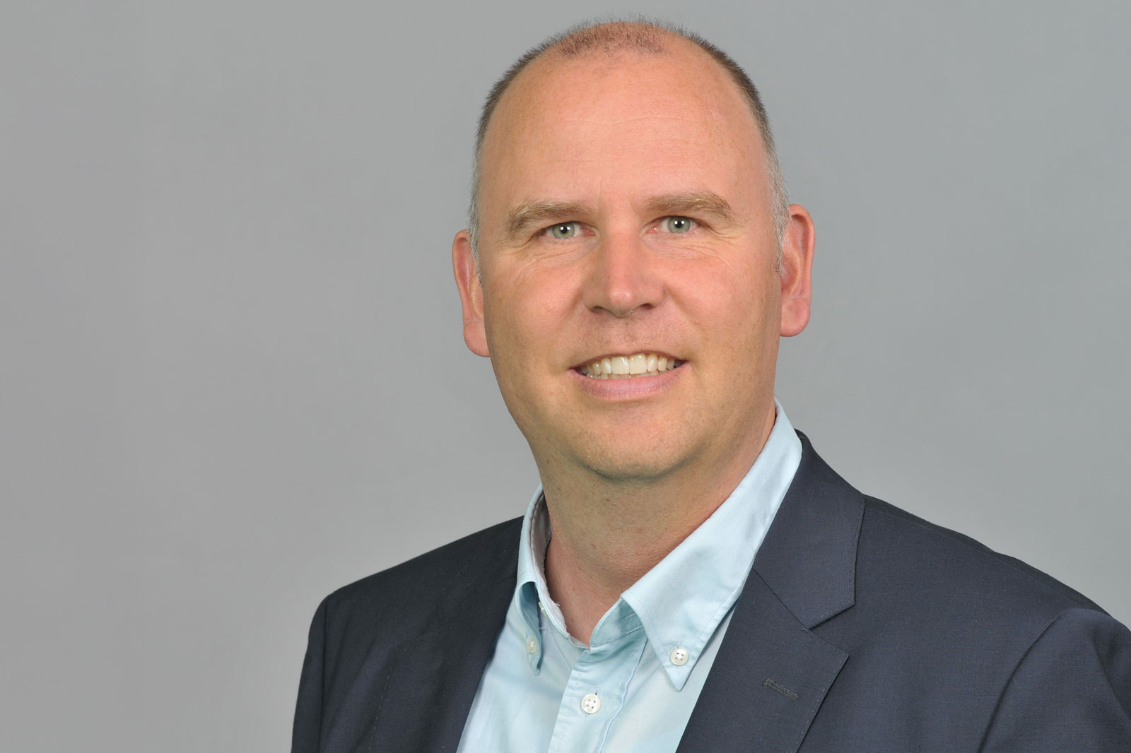 A man in a dark navy blazer and light blue shirt stands against a neutral gray background. Portrait of Philipp Biallass.
