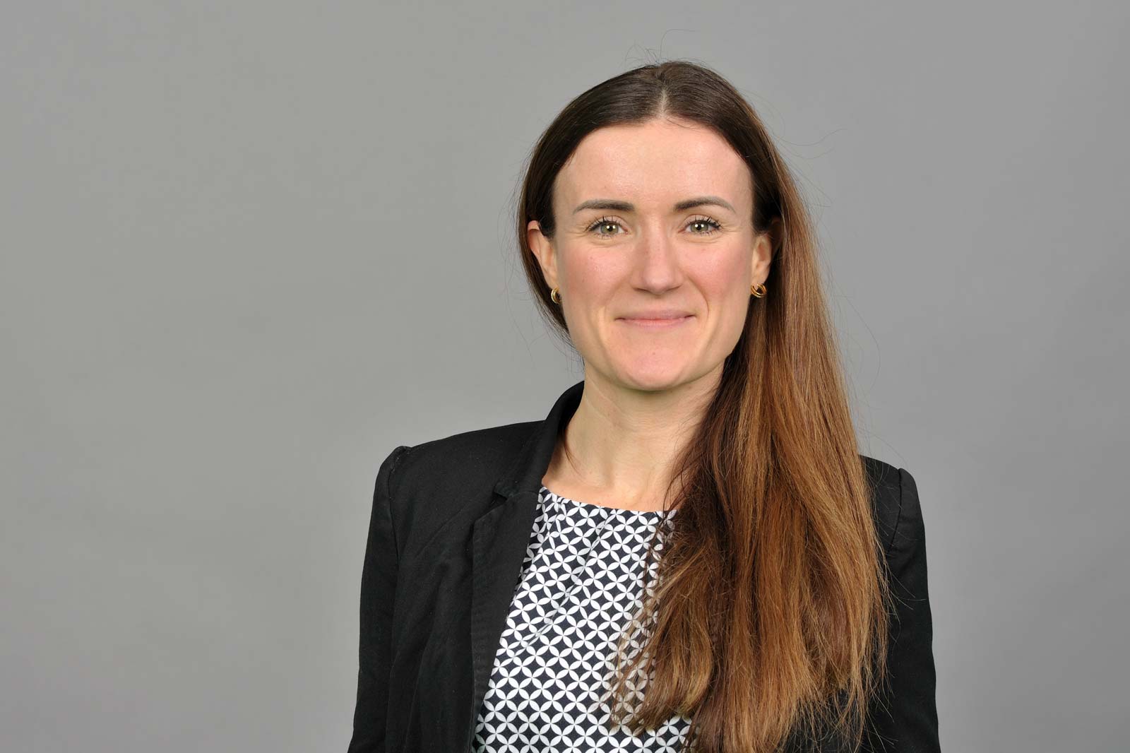 A woman with long brown hair is wearing a black blazer and a patterned top against a grey background. Portrait of Svenja Behr.