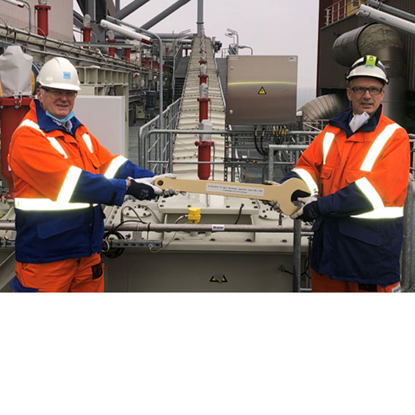 Two workers in protective clothing hold a large wrench on an industrial site.