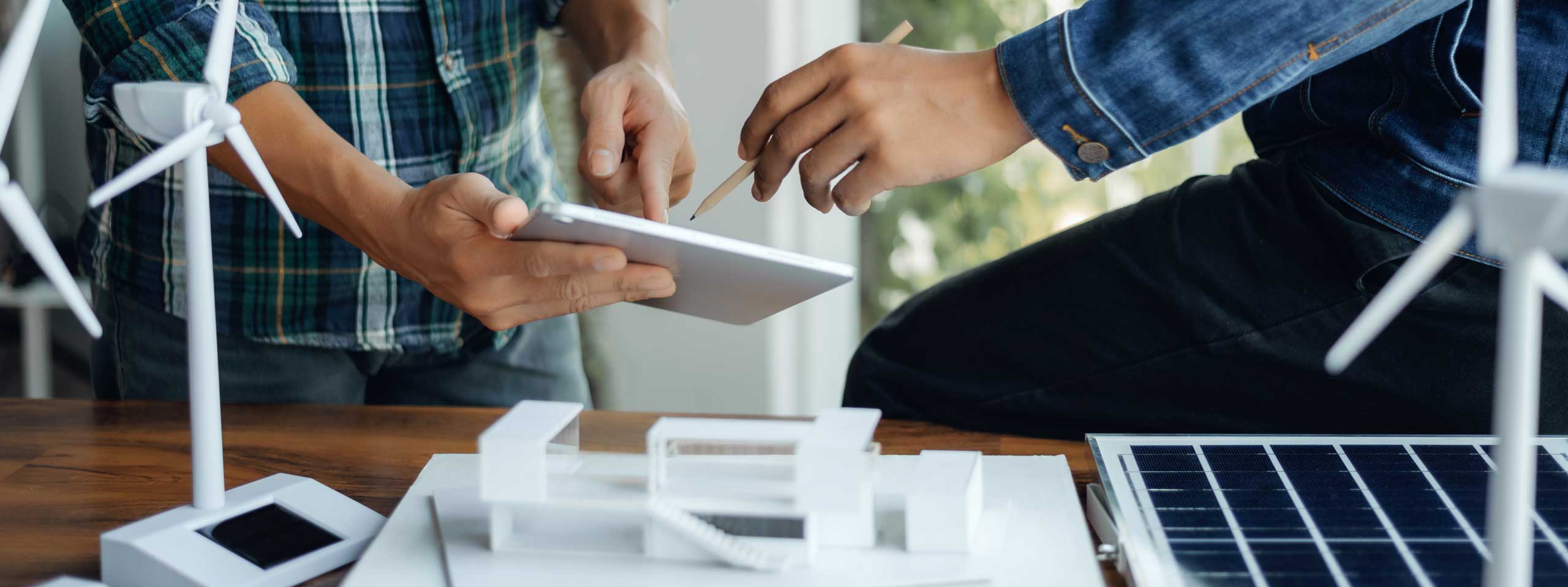 Zwei Personen arbeiten zusammen mit einem Tablet, Modell von Windkraftanlagen und Solarpanelen auf einem Tisch.