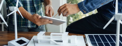 Zwei Personen arbeiten zusammen mit einem Tablet, Modell von Windkraftanlagen und Solarpanelen auf einem Tisch.