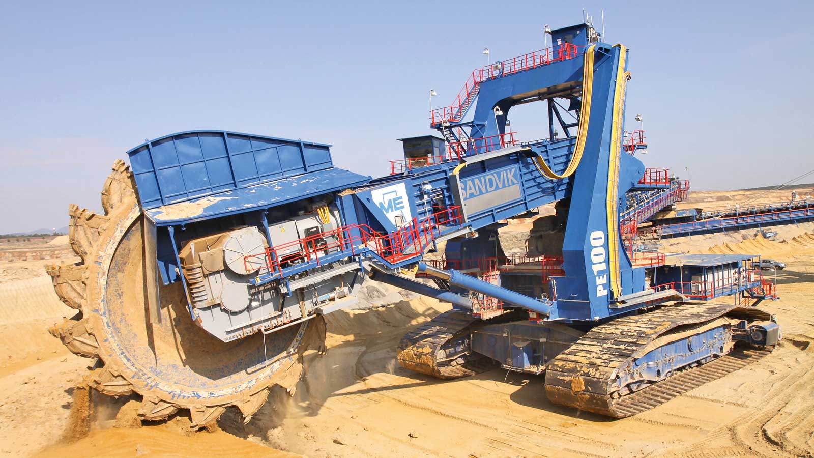 A large blue and grey continuous miner is excavating dirt at a mining site under a clear sky.