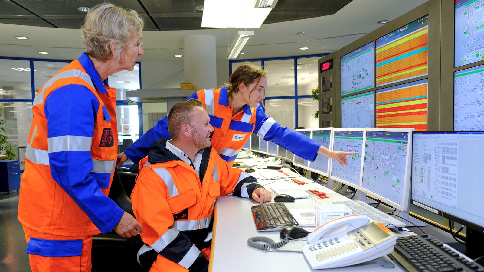 Three workers in safety gear collaborate at a control room with multiple screens displaying data and system statuses.