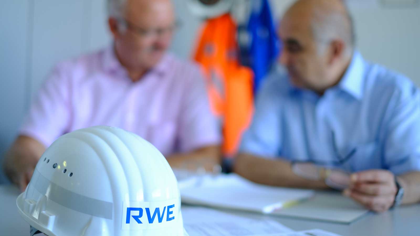 A white safety helmet with the RWE logo rests on a table beside two individuals discussing documents.