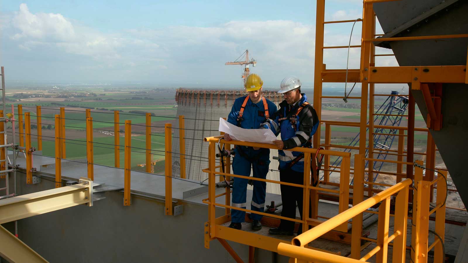Two construction workers review plans on a building site, surrounded by scaffolding and an expansive landscape.
