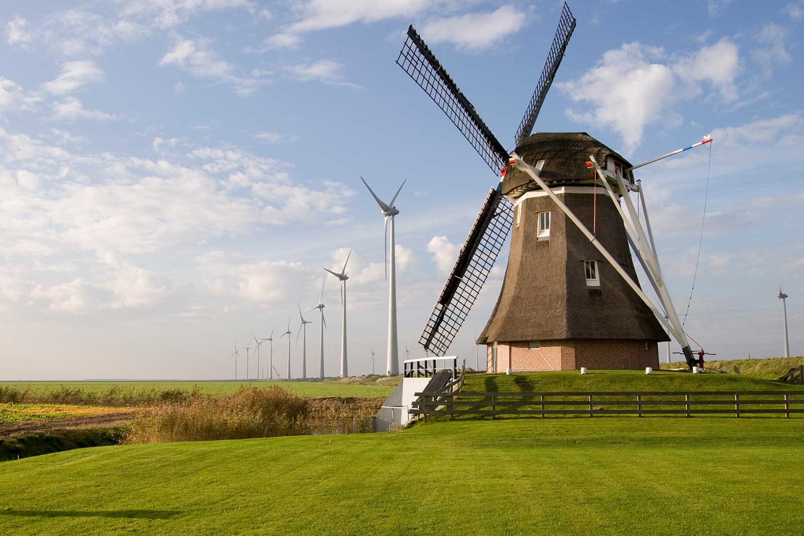 A traditional windmill stands beside modern wind turbines under a blue sky with clouds.