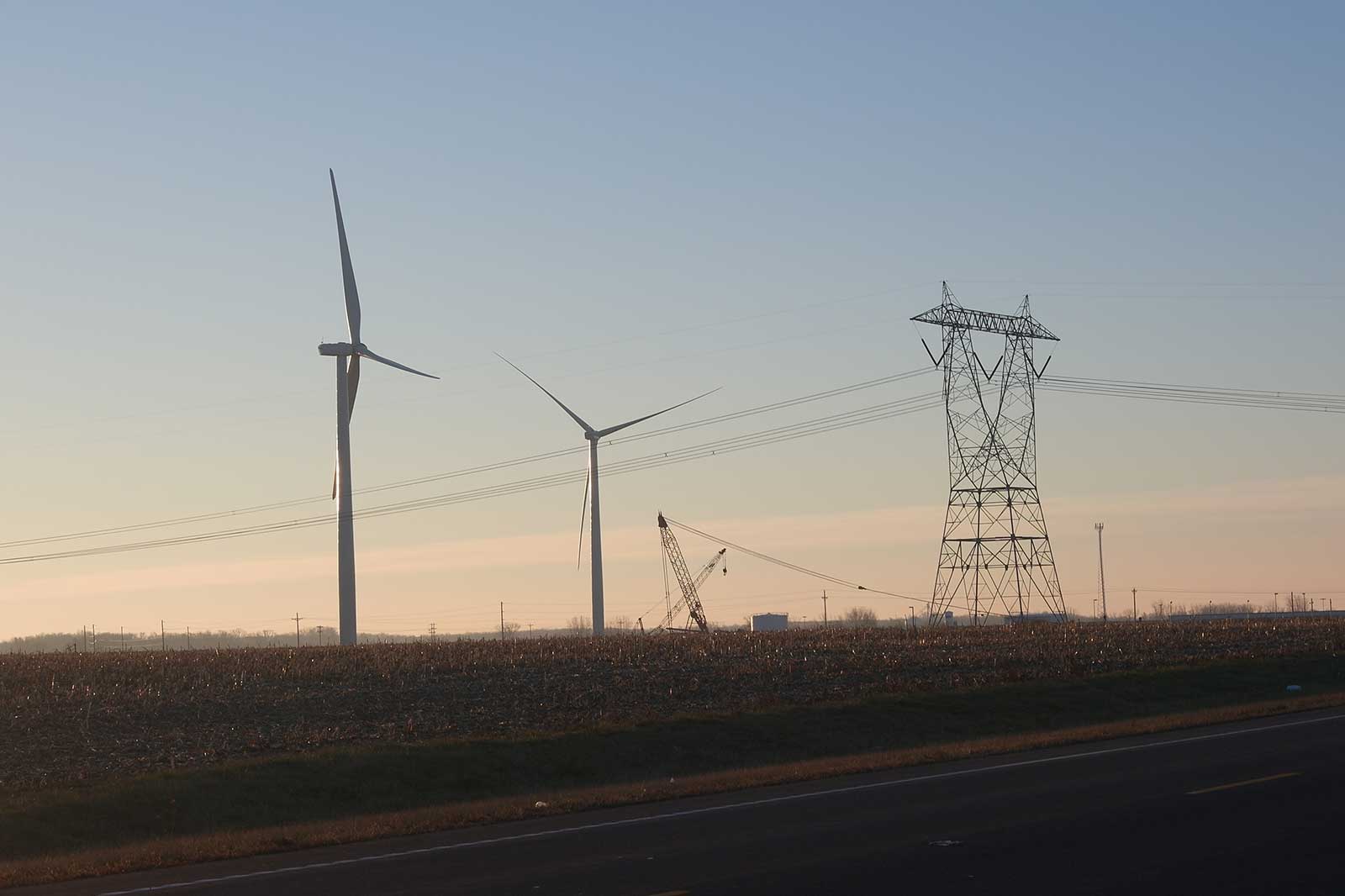 Wind turbines and power pylons in a rural setting at dawn, with a view of the road in the foreground.