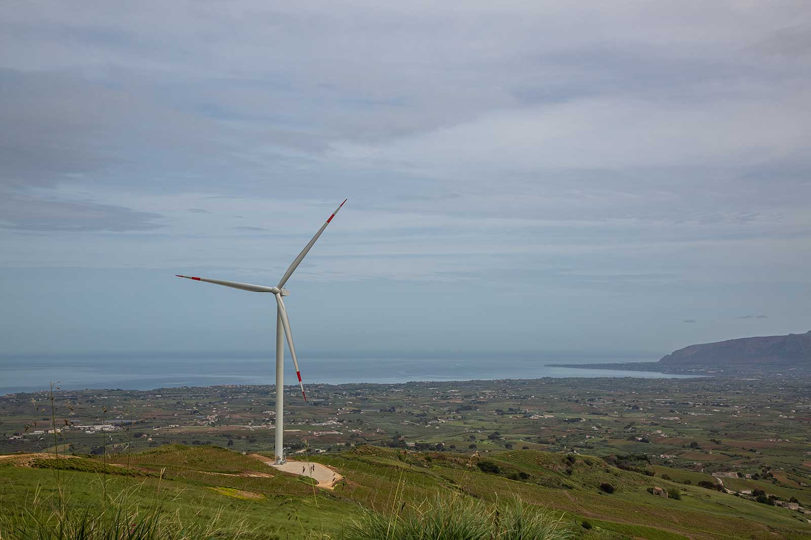 A wind turbine stands on a hill overlooking a green valley and the sea in the background under a cloudy sky.
