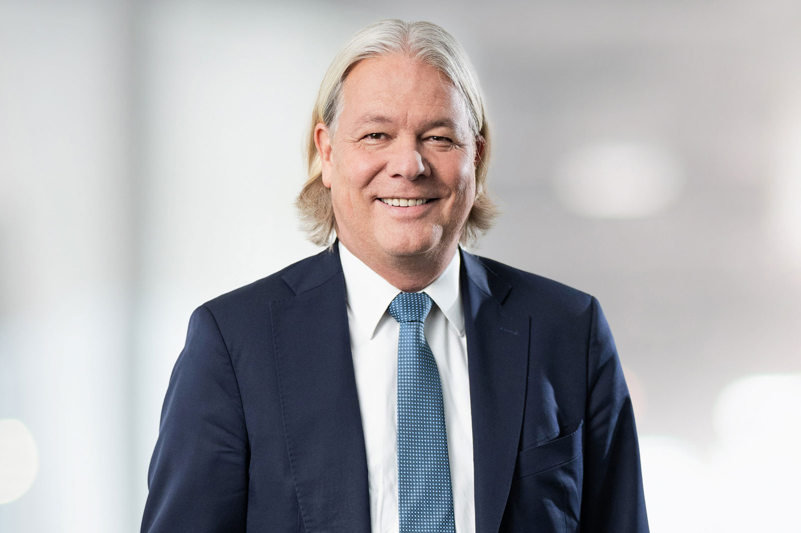 A man in a suit with a tie stands in front of a blurred, bright background.
