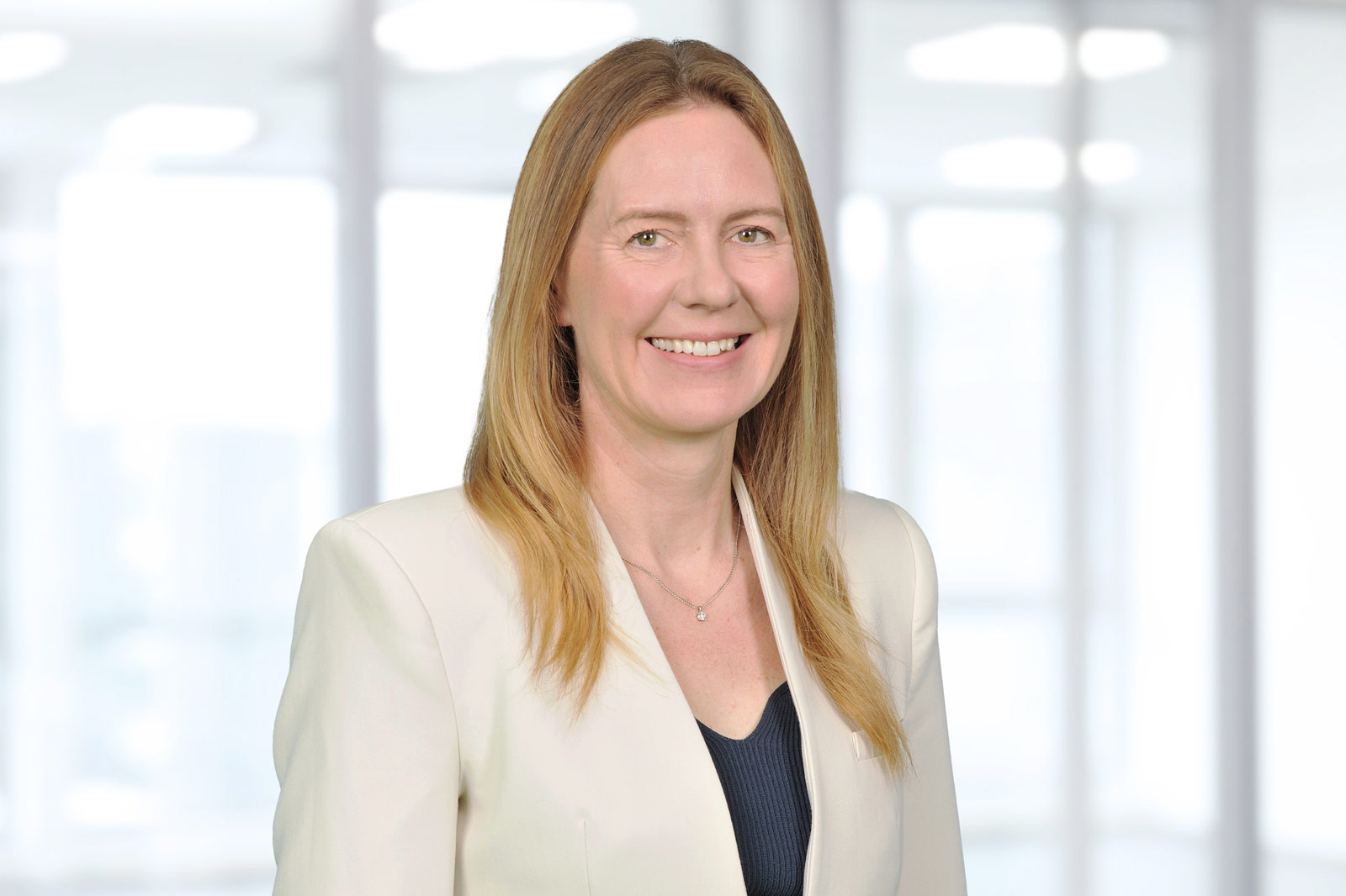 A person stands with crossed arms against a blurred, bright background. They are wearing a white blazer with black accents.