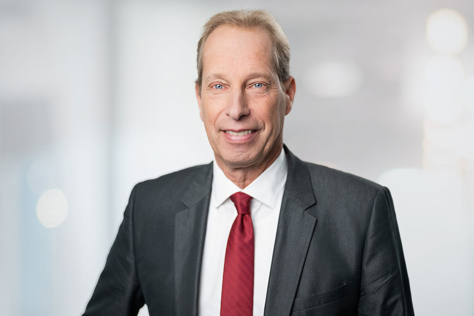 Ein Mann in einem Anzug mit roter Krawatte steht vor einem verschwommenen Hintergrund.