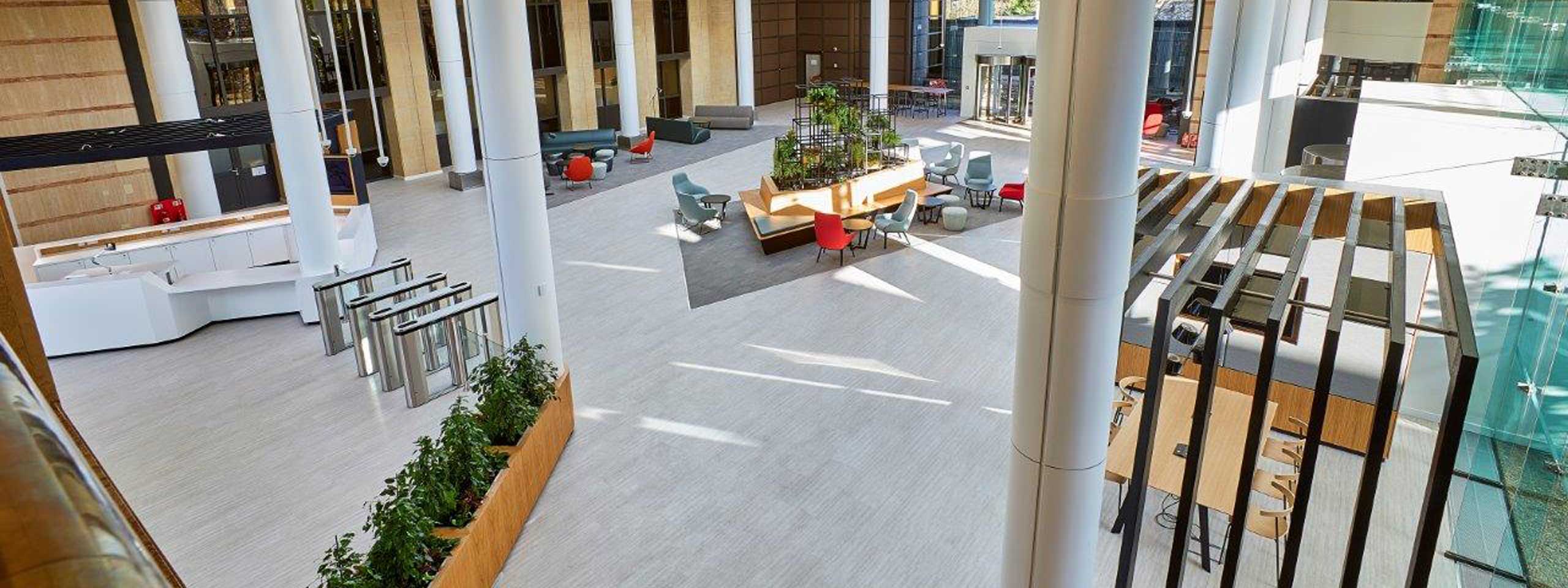 A modern lobby area featuring seating, plants, and a reception desk. Large windows provide natural light.