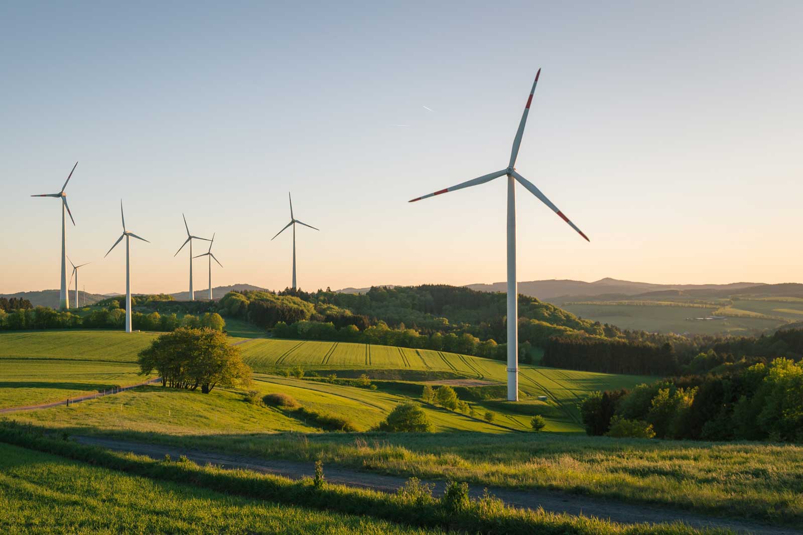 Fünf Windkraftanlagen stehen auf einem grünen Hügel unter einem klaren Himmel bei Sonnenuntergang.