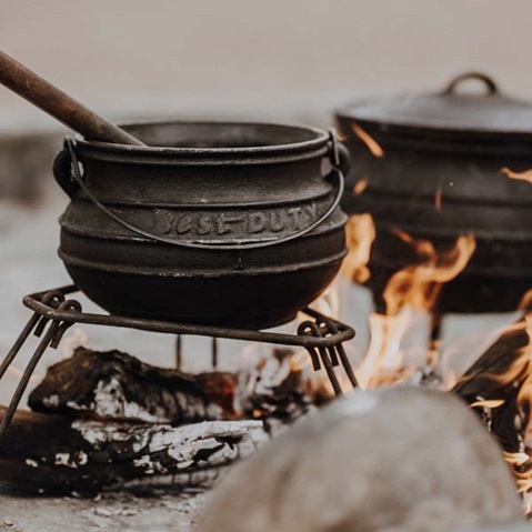 A cast iron pot over an open fire. The pot has a wooden spoon and is supported by a sturdy stand.