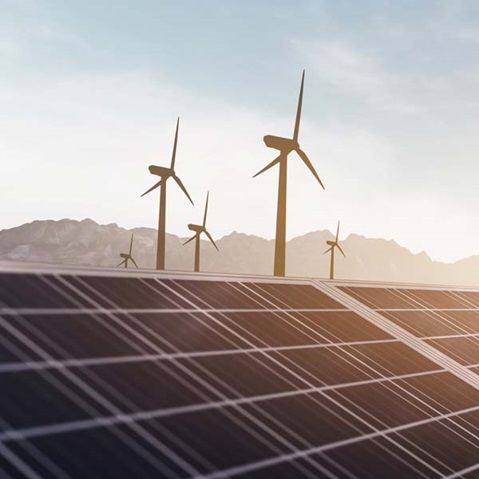A solar panel field with wind turbines in the background against a mountain and clear sky horizon.