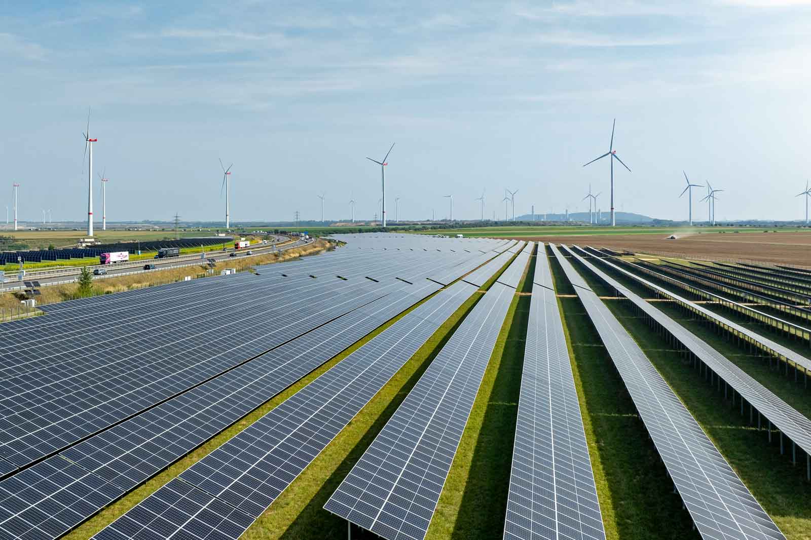 Eine weite Fläche mit Solarpanelen, umgeben von Windkraftanlagen und einer Straße im Hintergrund.
