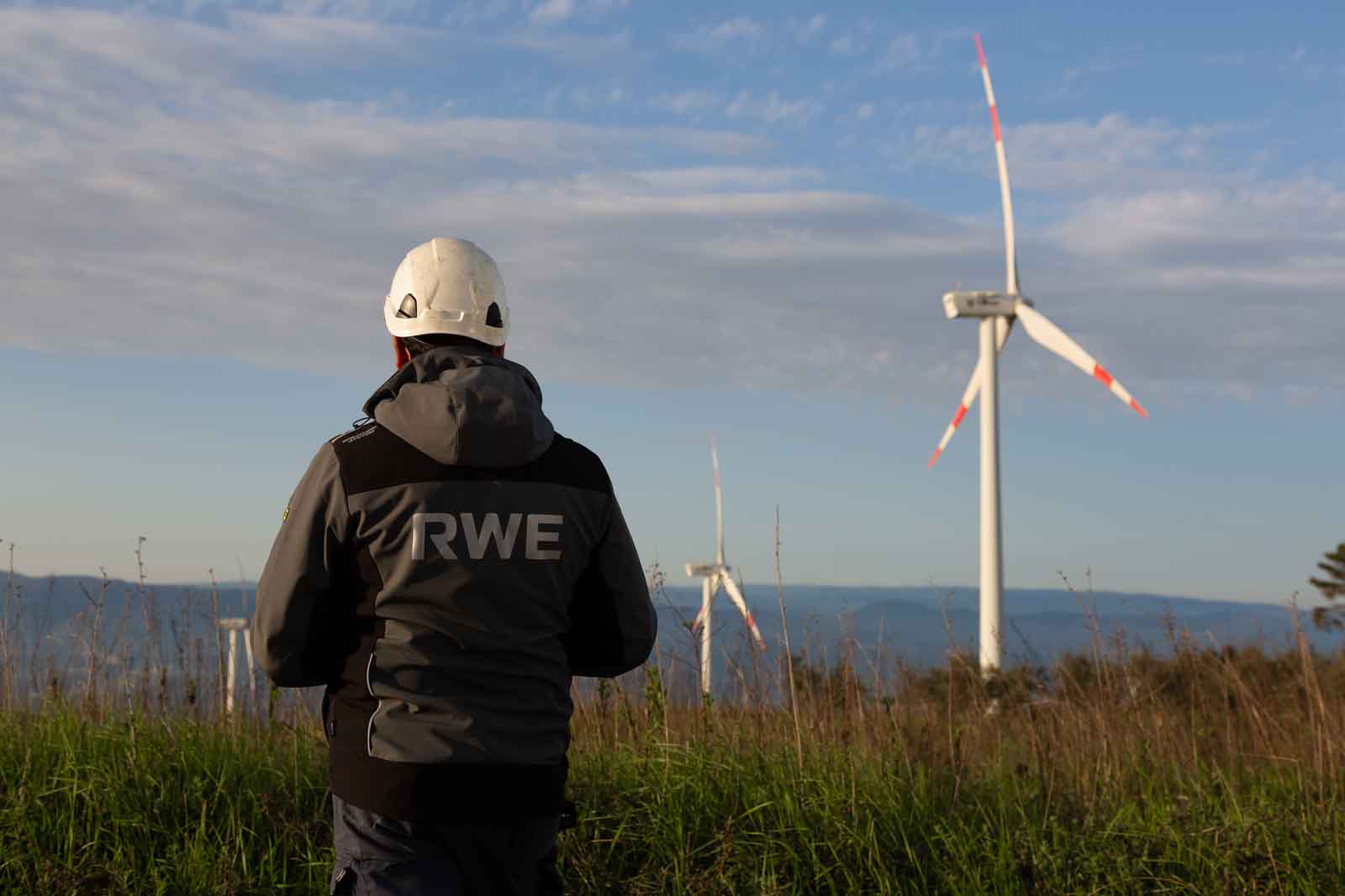 Ein Techniker mit einem Helmut steht vor Windkraftanlagen, umgeben von Gras und einer sanften Hügellandschaft unter blauem Himmel.
