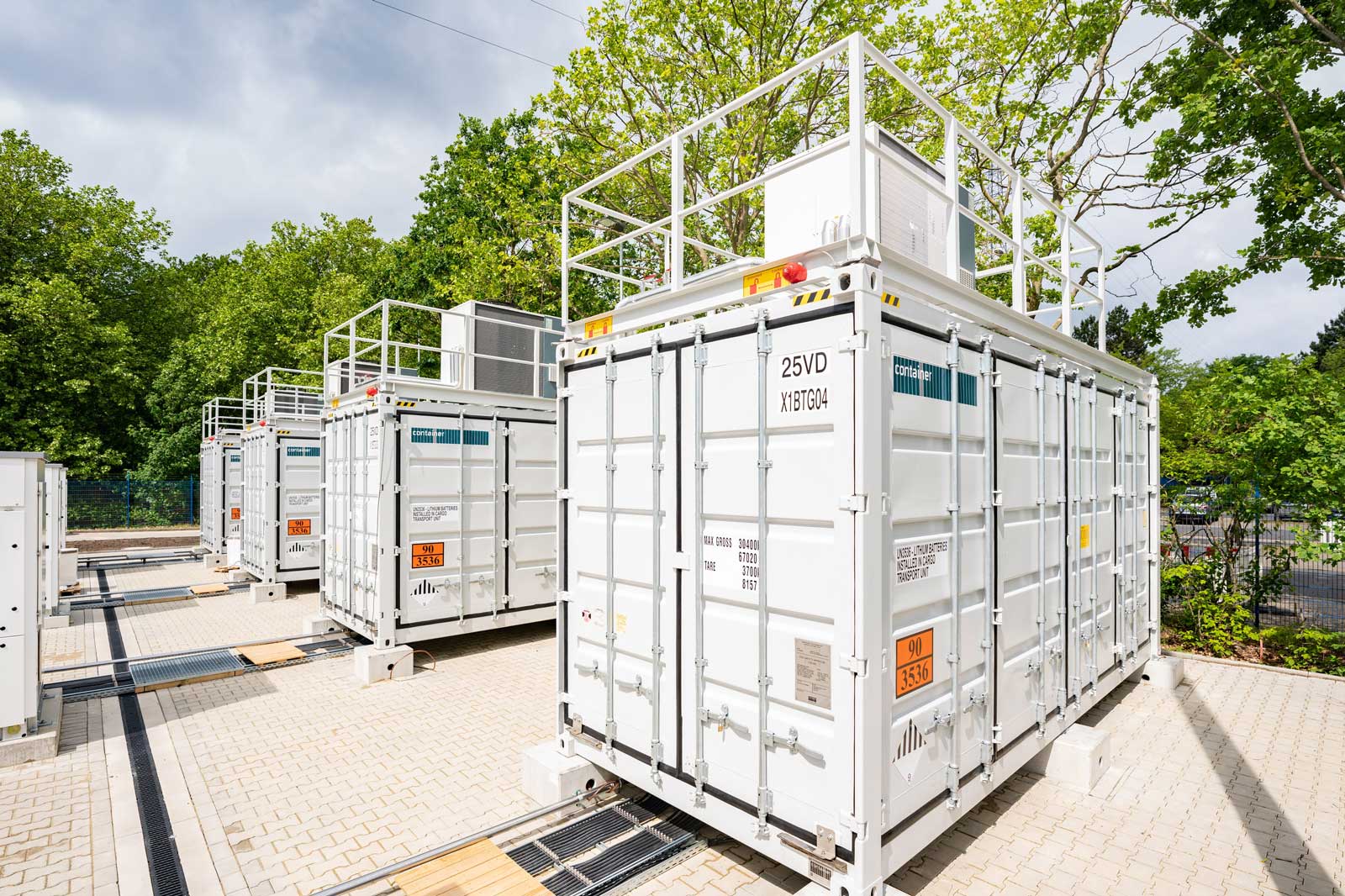 Reihe von weißen Lagercontainern mit Leitern in einem grünen Außenbereich unter einem bewölkten Himmel.