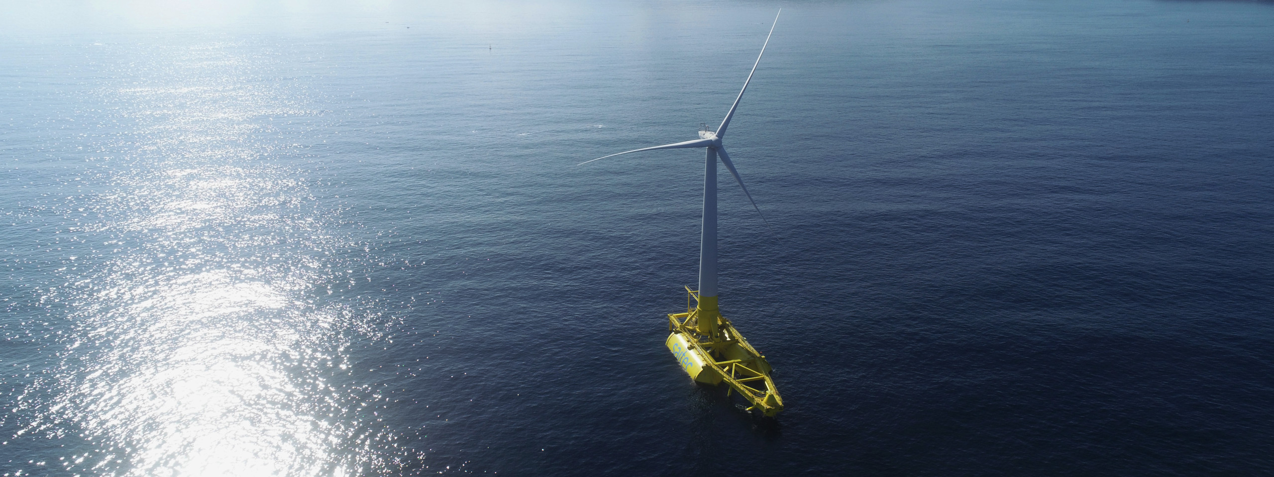 A yellow floating wind turbine stands still in the blue sea, as sunlight reflects off the water's surface.