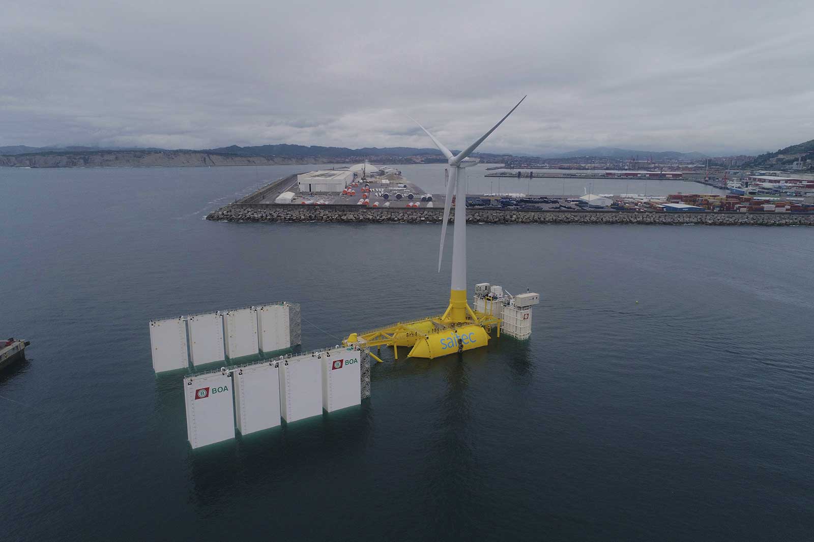 A floating wind turbine with a yellow base and white floats in calm water. A harbour is visible in the background.