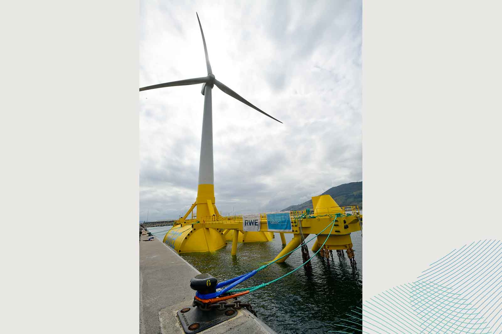 A wind turbine towers above a yellow platform in the water, set against a grey sky.