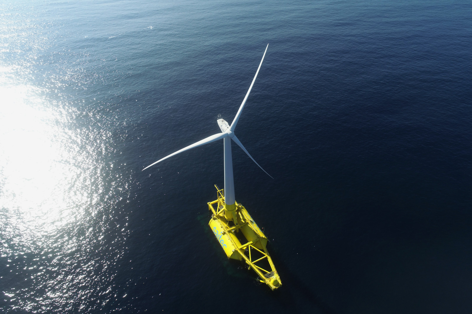 A yellow offshore wind turbine stands on the water, with its blades rotating in the wind.