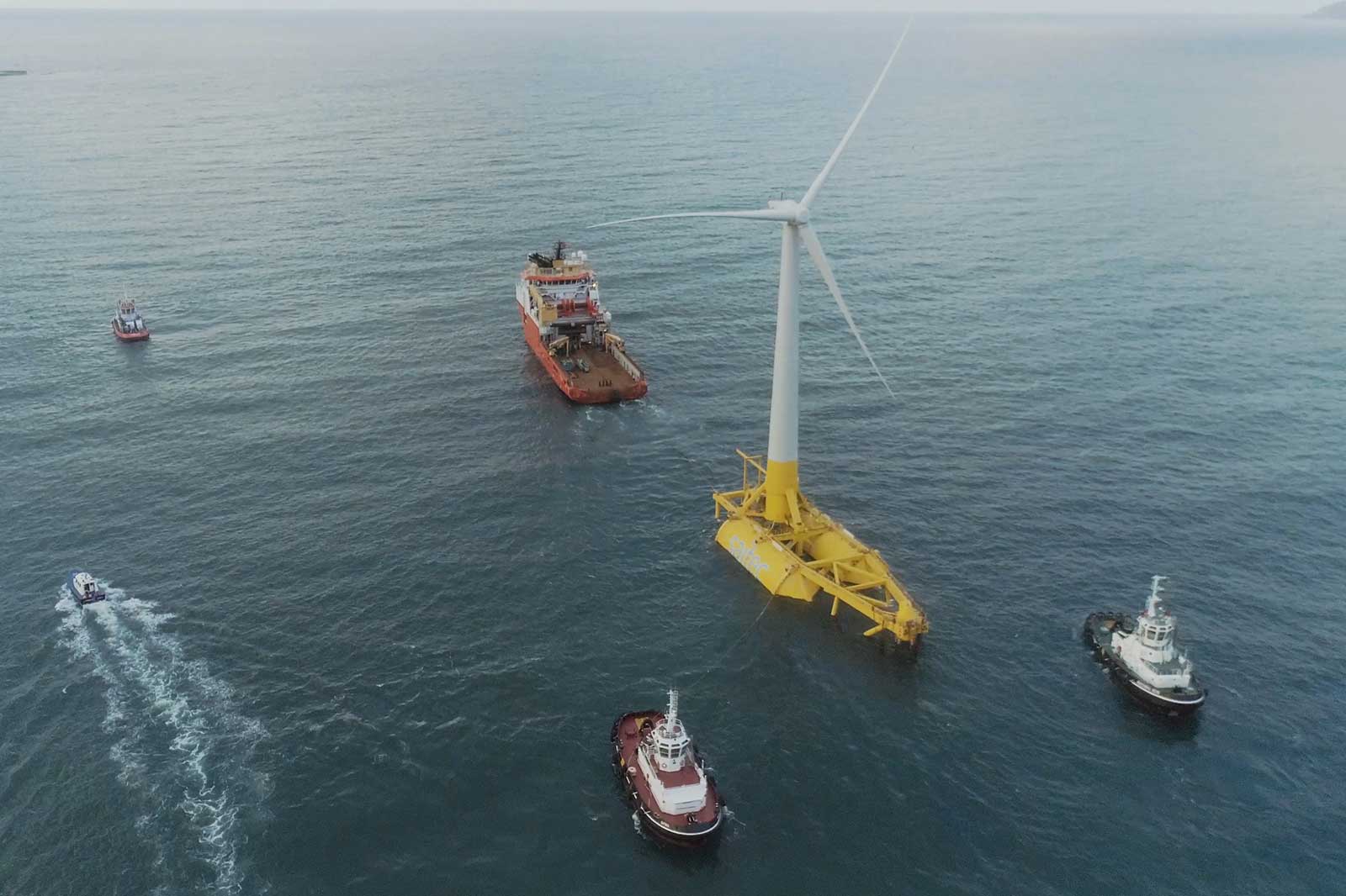 An offshore wind turbine stands in the water, surrounded by several tugboats and a service vessel.