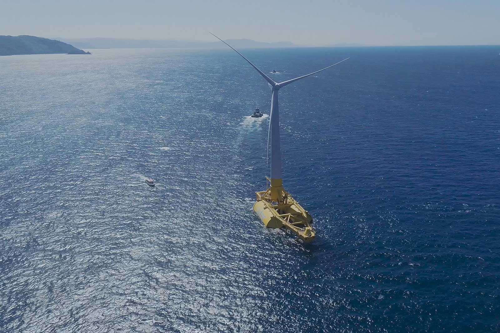 An aerial view of a yellow offshore wind turbine in the ocean, with boats nearby and mountains in the background.