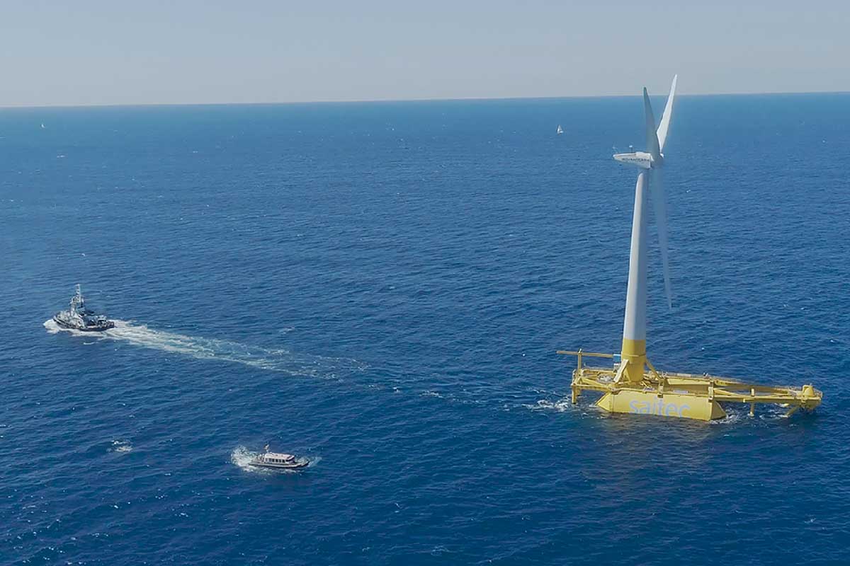 An offshore wind turbine stands in blue water, while a tugboat and a small vessel navigate nearby.