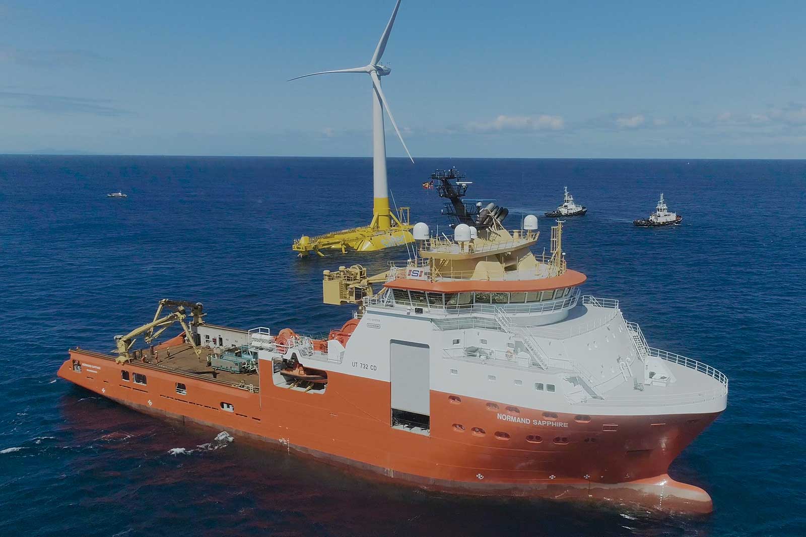 An orange ship, the Normand Sapphire, conducts operations near a wind turbine in the ocean.