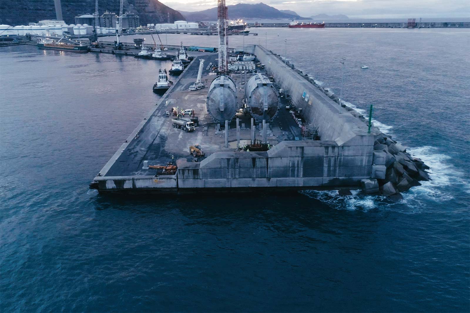 A harbour area featuring a concrete pier and two large cylindrical tanks on the dock. Ships are visible in the background.
