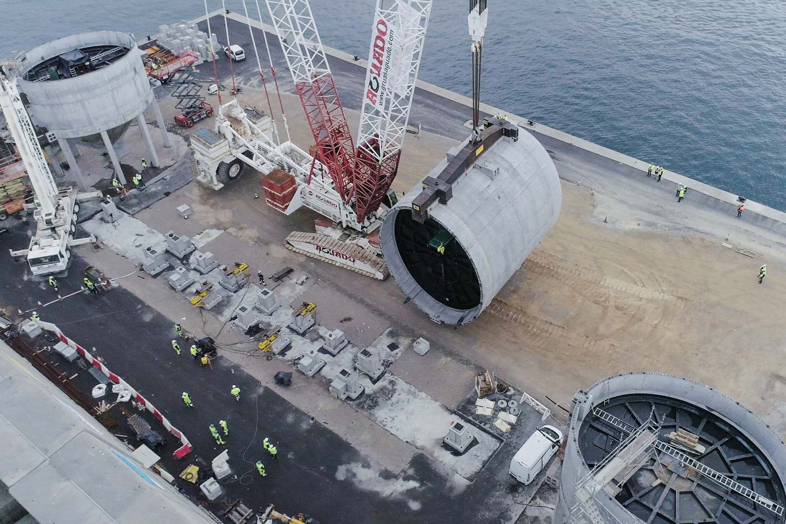 Construction work at a port facility with cranes, workers, and large concrete cylinders.