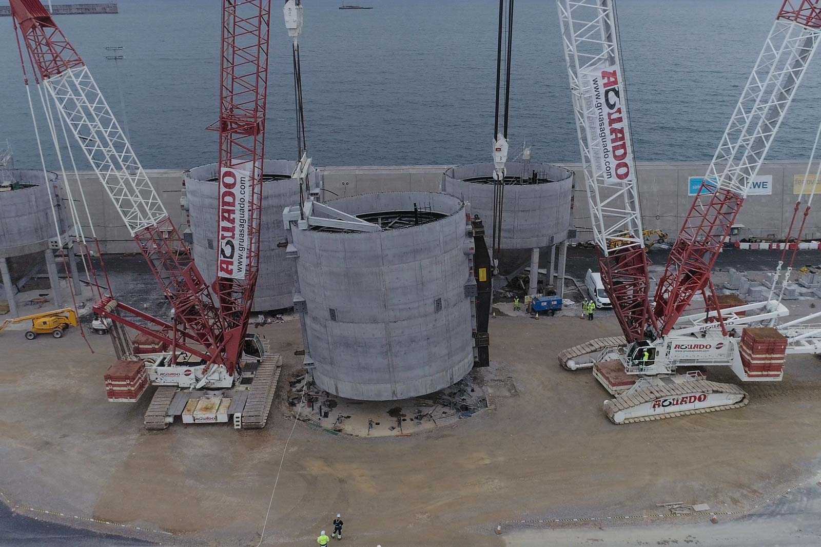 A construction site by the sea with large cranes working on concrete cylinders. Workers are visible.