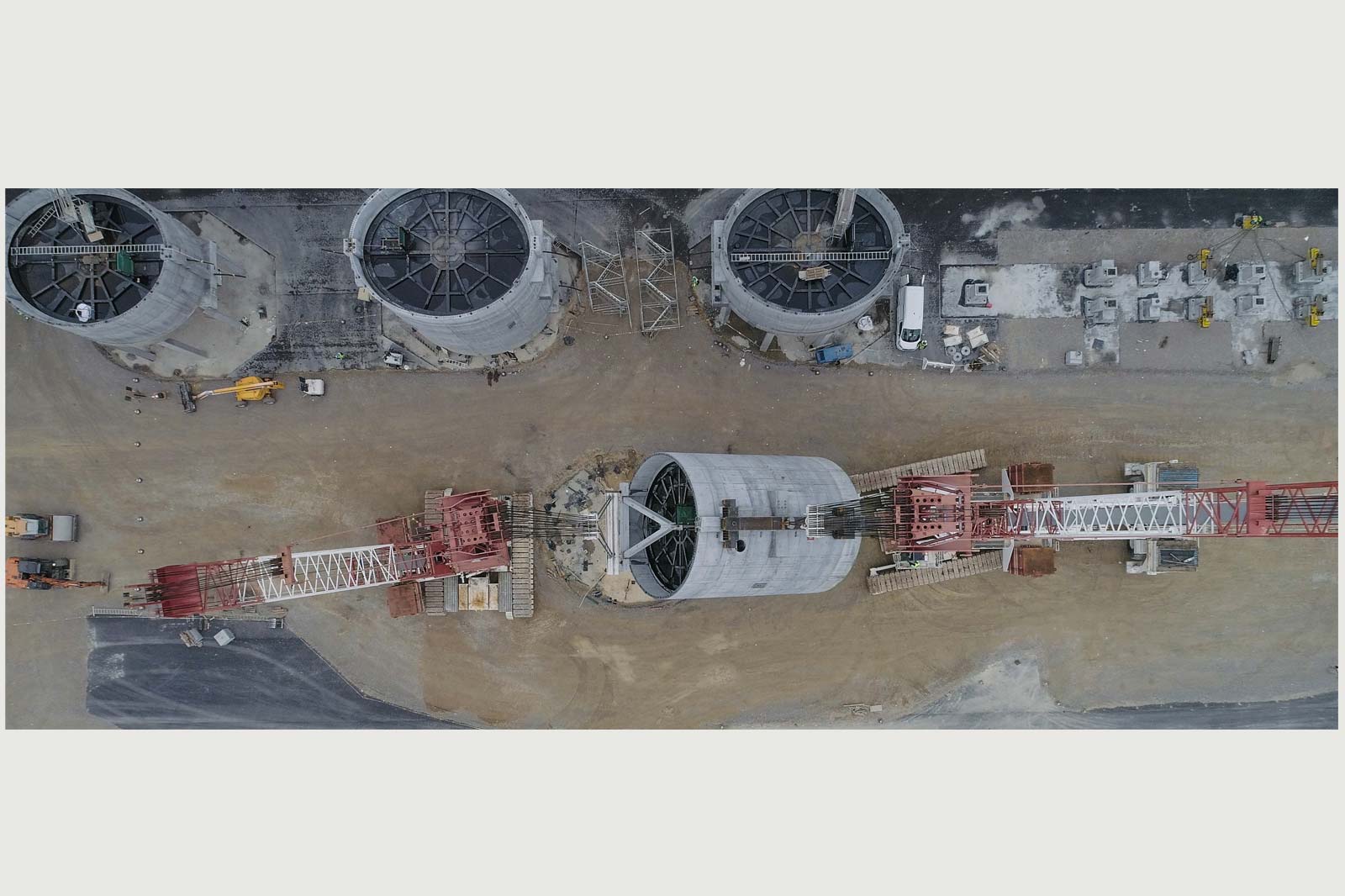 A construction site featuring three large cylindrical structures with a crane in the centre. Materials and machinery are visible.