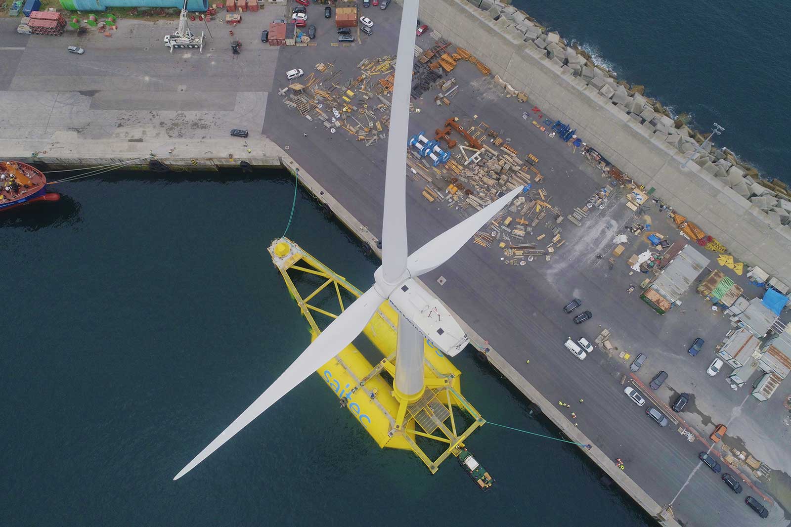 Eine Offshore-Windkraftanlage auf einem gelben Ponton vor dem Hafen. Fahrzeuge und Materialien sind im Hintergrund sichtbar.