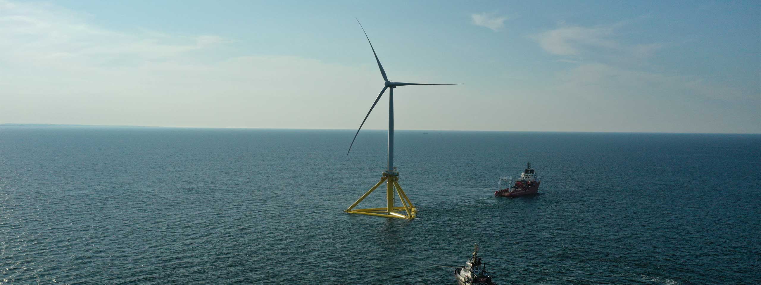 A single offshore wind turbine stands in the water, surrounded by gentle waves, with a ship in the background.