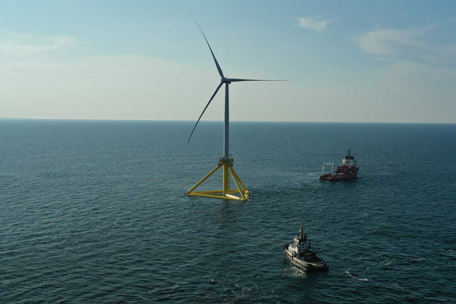 An offshore wind turbine stands in the sea, while a boat sails nearby.