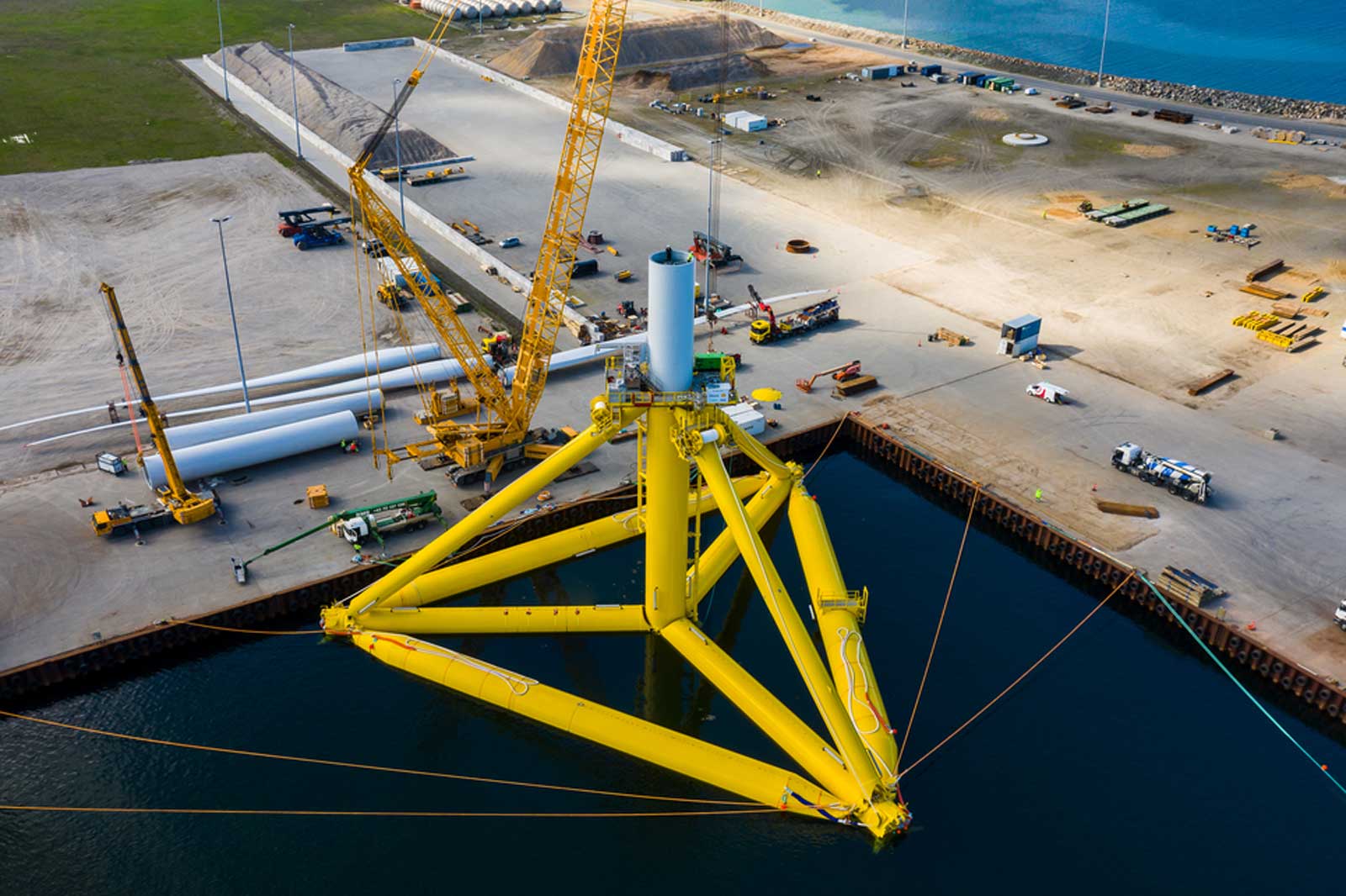 A yellow wind turbine foundation in water, surrounded by construction equipment and pipes on land.