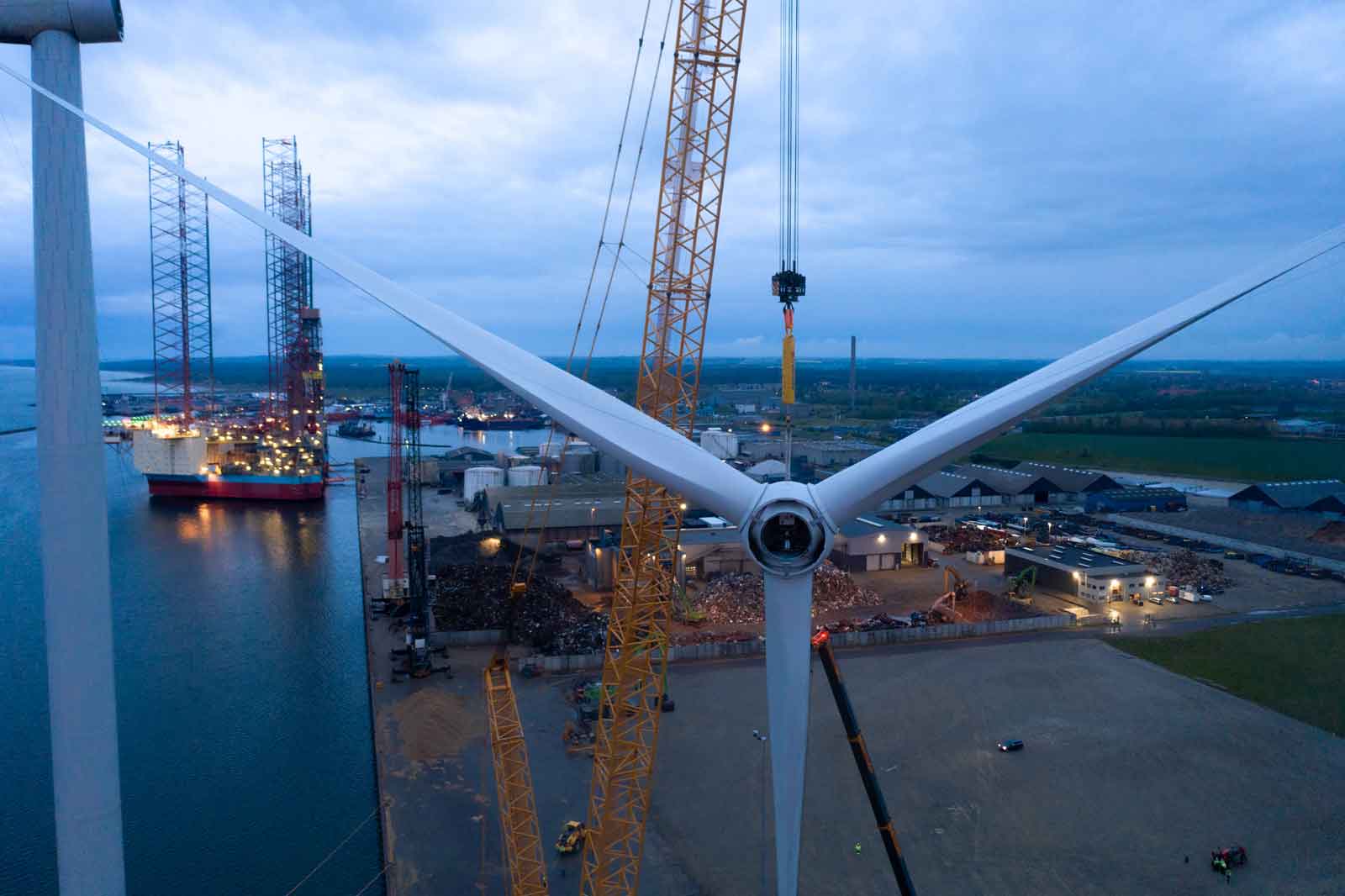 View of a wind turbine construction site with cranes and ships in the harbour under a cloudy sky.