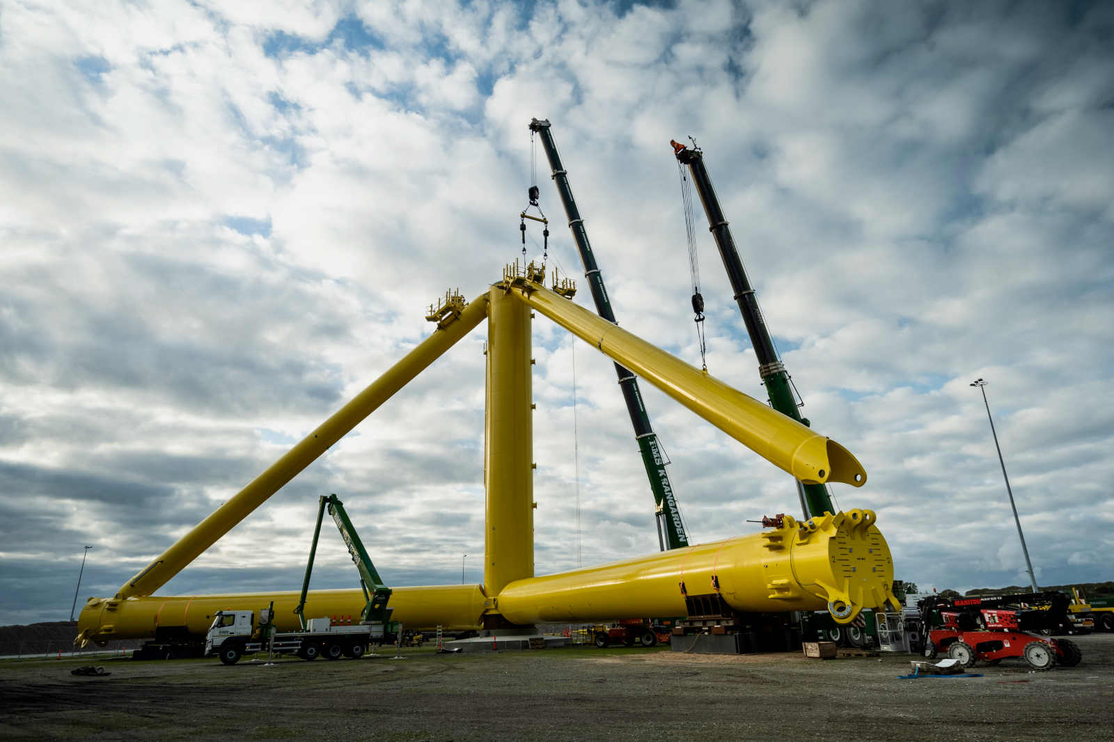 Large yellow construction components are being lifted by cranes at a construction site, set against a cloudy sky.