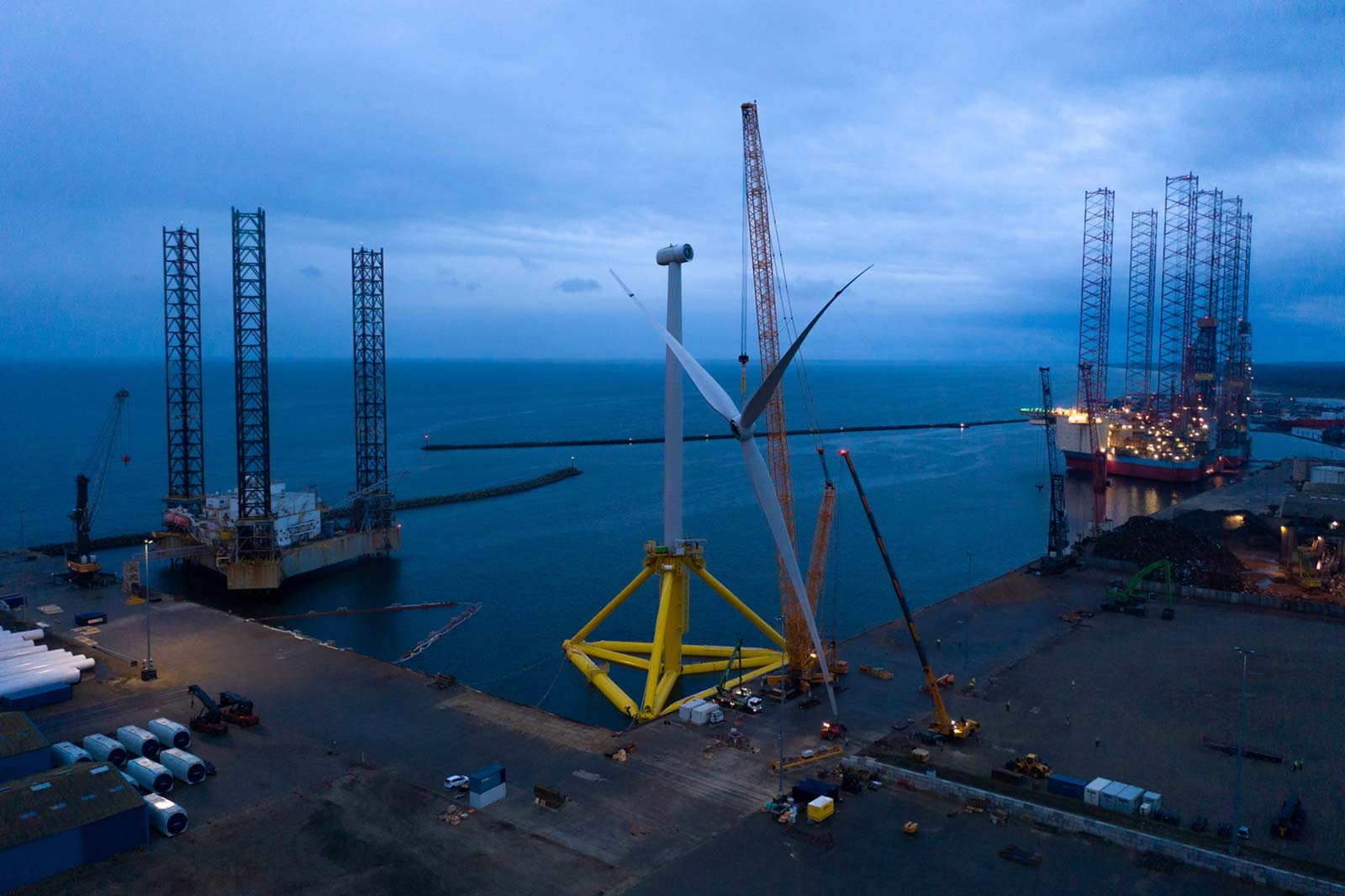 An offshore wind farm with a large turbine, cranes, and an ocean backdrop at dusk.