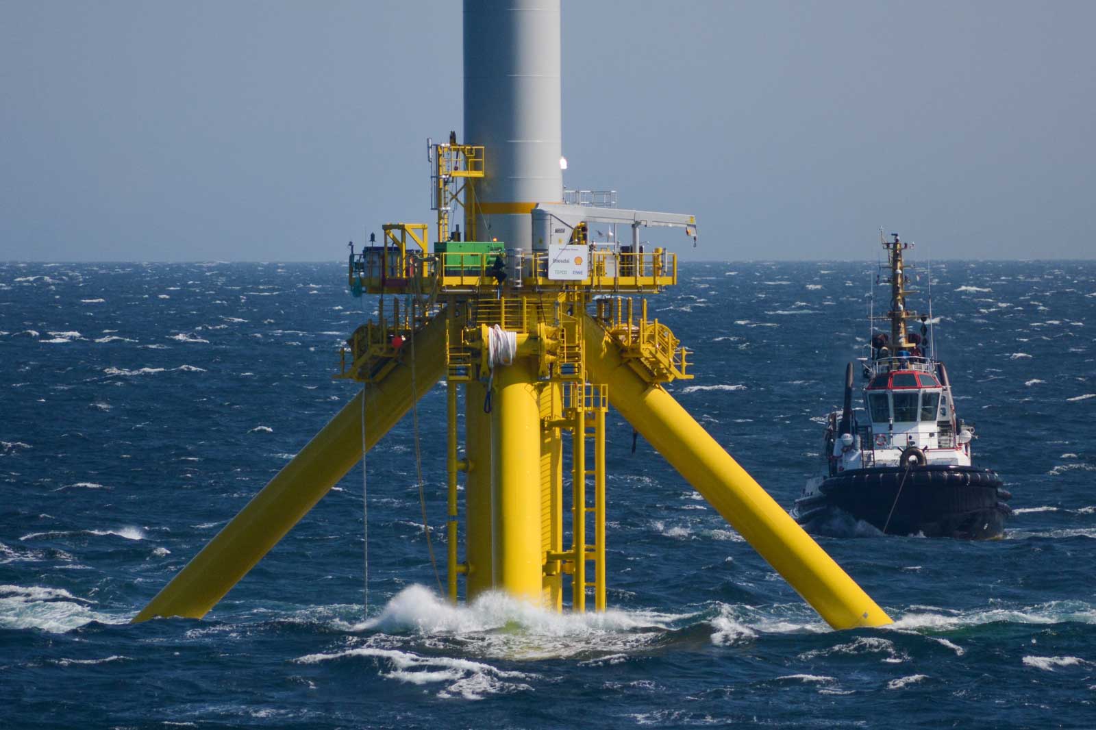 A yellow offshore wind turbine tower stands in the sea, with a tugboat nearby.