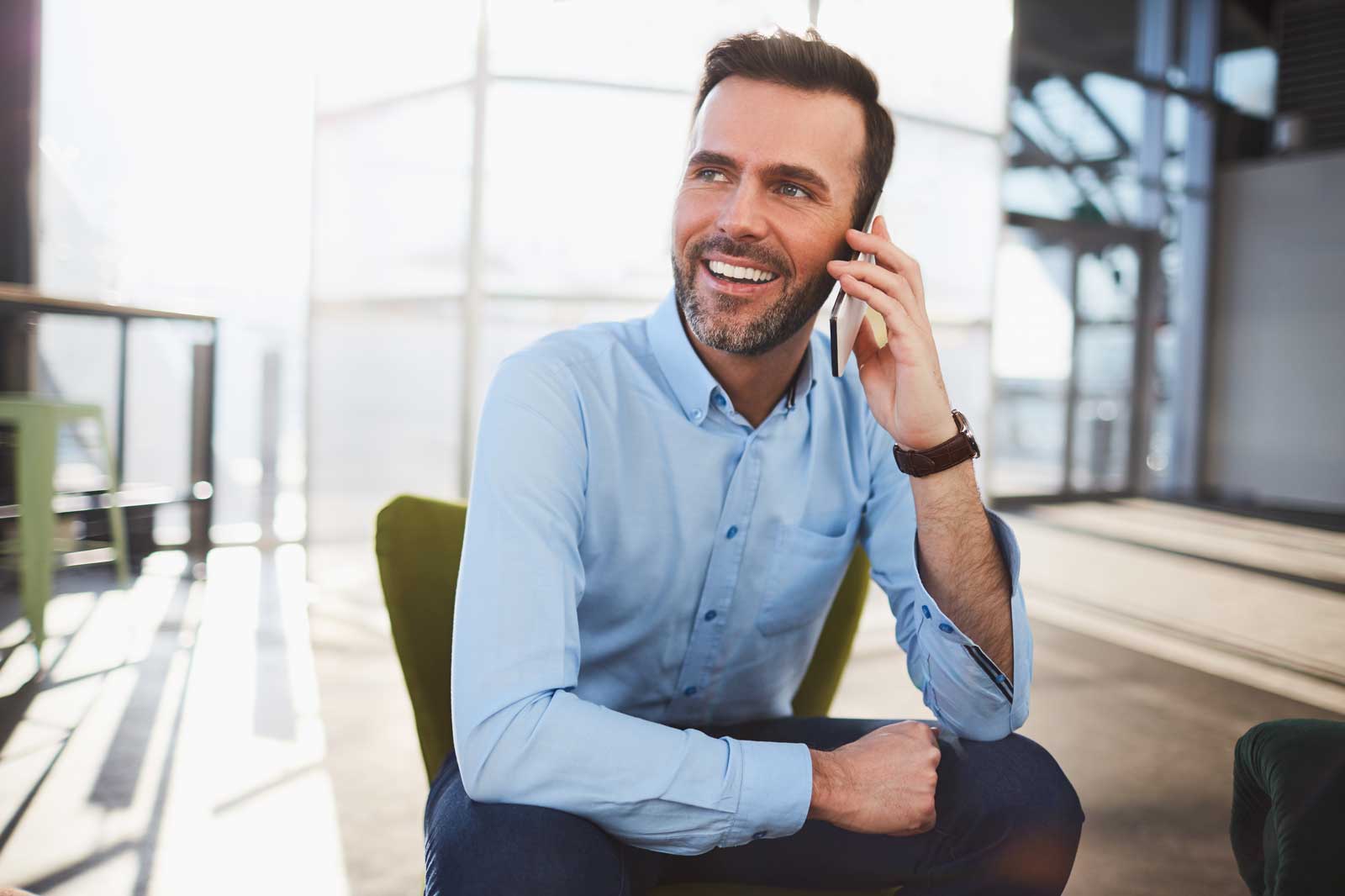 Ein Mann in einem hellblauen Hemd sitzt auf einem Stuhl und telefoniert in einer modernen Büroumgebung.