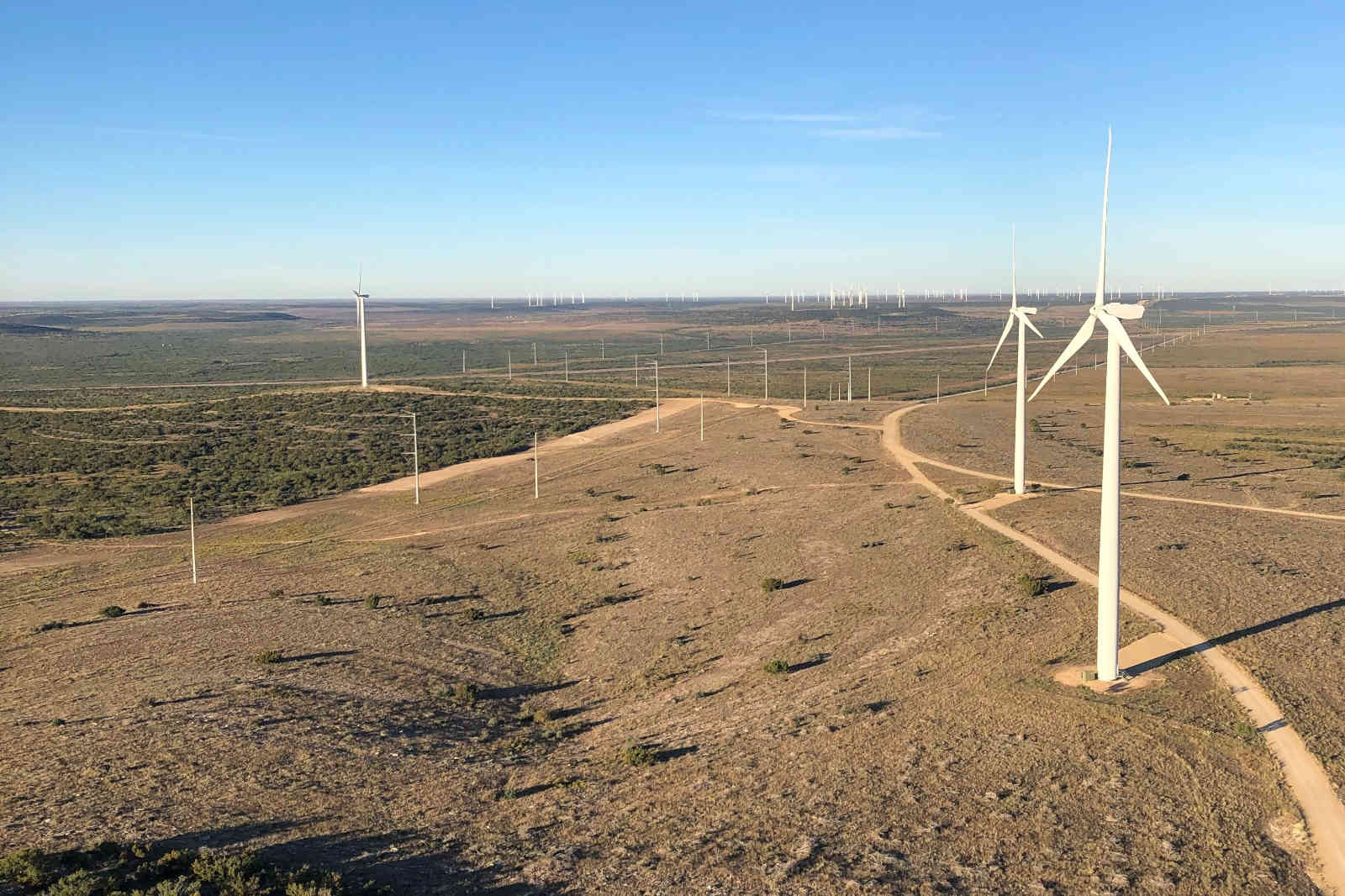 Aerial view of wind turbines on a vast plain under a clear blue sky, with dirt paths and green vegetation in the foreground.