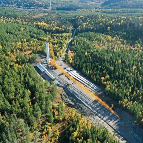 Luftaufnahme eines bewaldeten Gebiets mit Windkraftanlagenkomponenten und einem Kran auf einer Baustelle.
