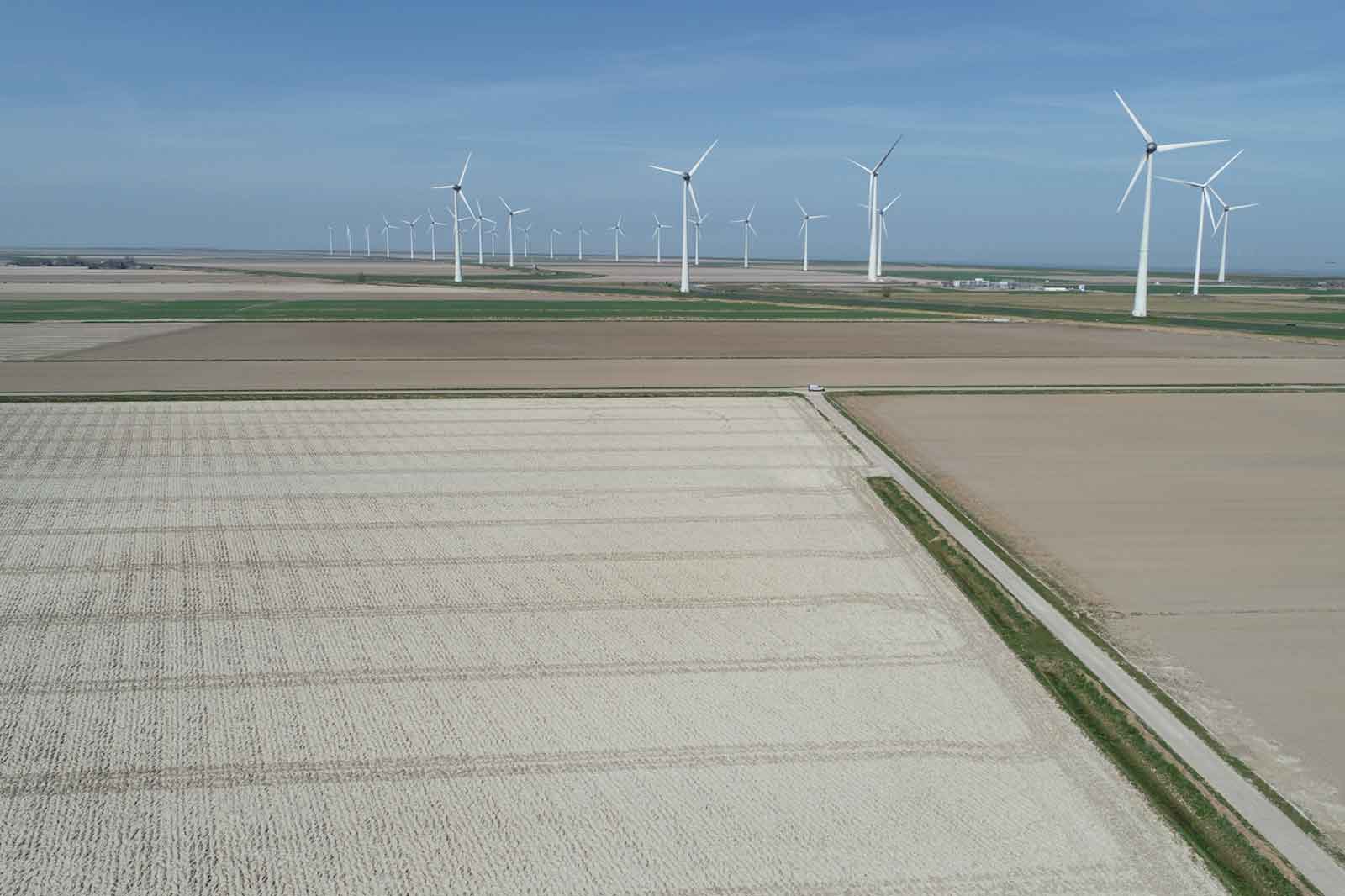 Luftaufnahme einer Windfarm mit mehreren Windturbinen über Feldern in verschiedenen Farben unter einem blauen Himmel.