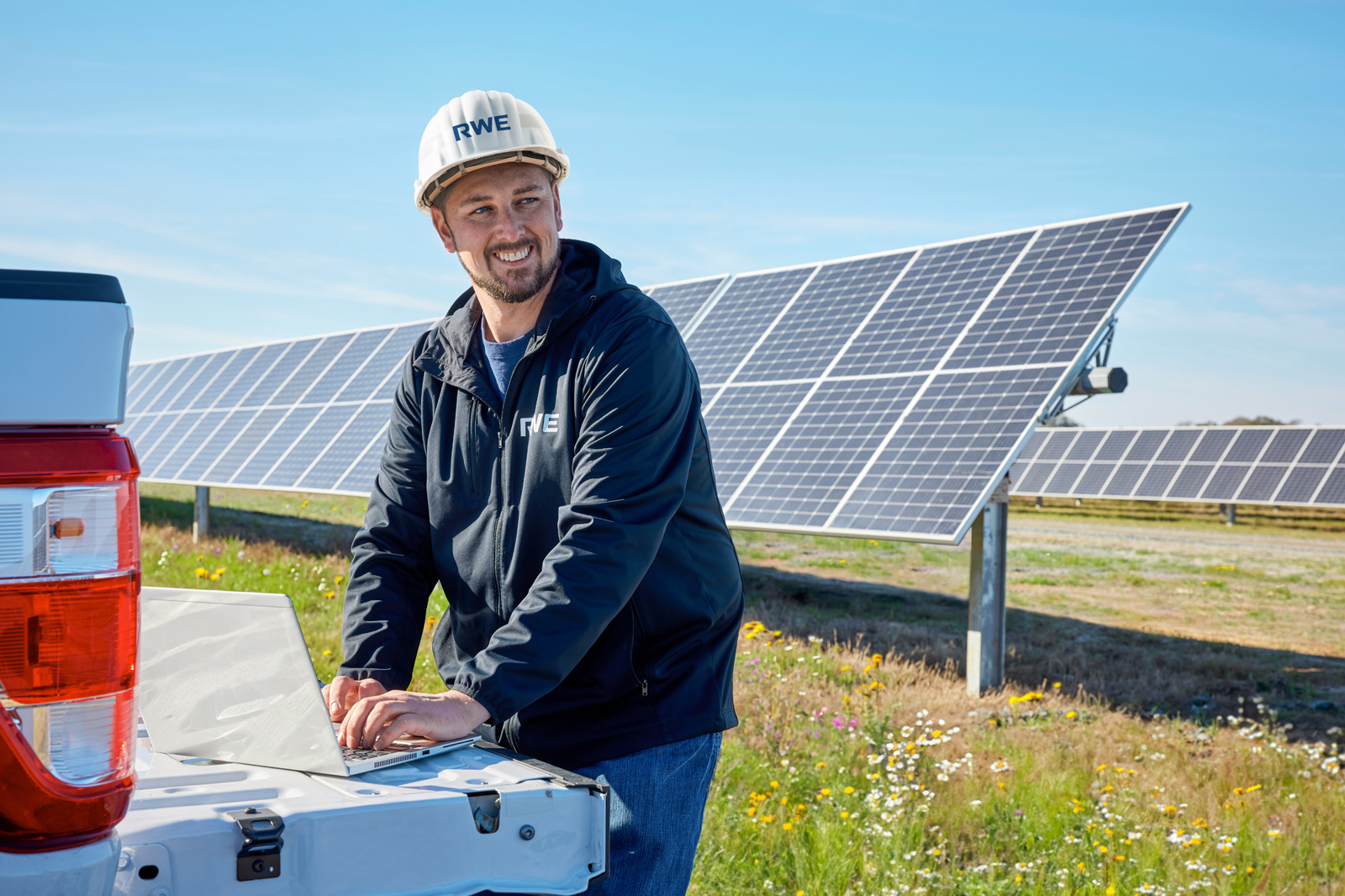 A worker in a hard hat uses a laptop on the tailgate of a truck near solar panels under a clear blue sky.