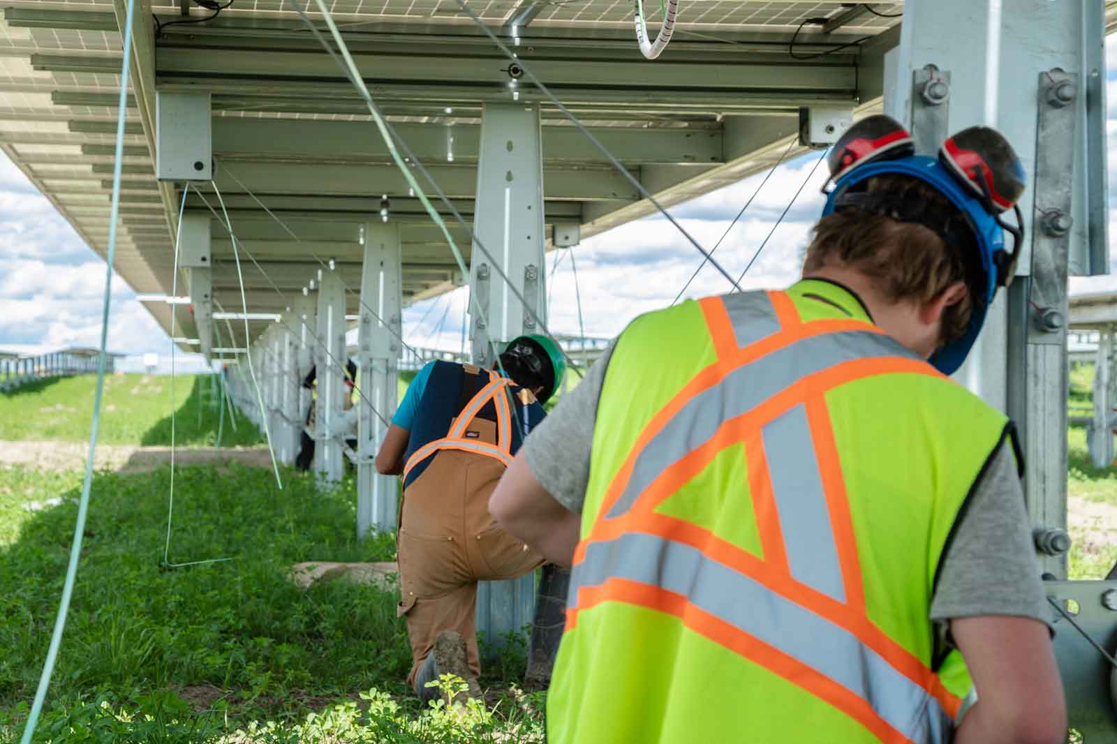 Zwei Arbeiter in reflektierenden Sicherheitswesten arbeiten unter Solarpanelen auf einer Wiese.