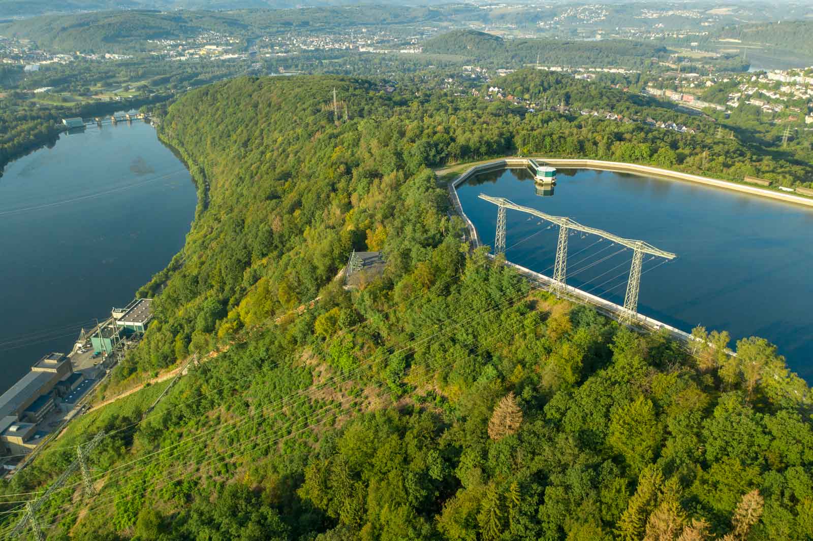 An aerial view of a wooded hill beside a lake, featuring electricity pylons and a water reservoir.