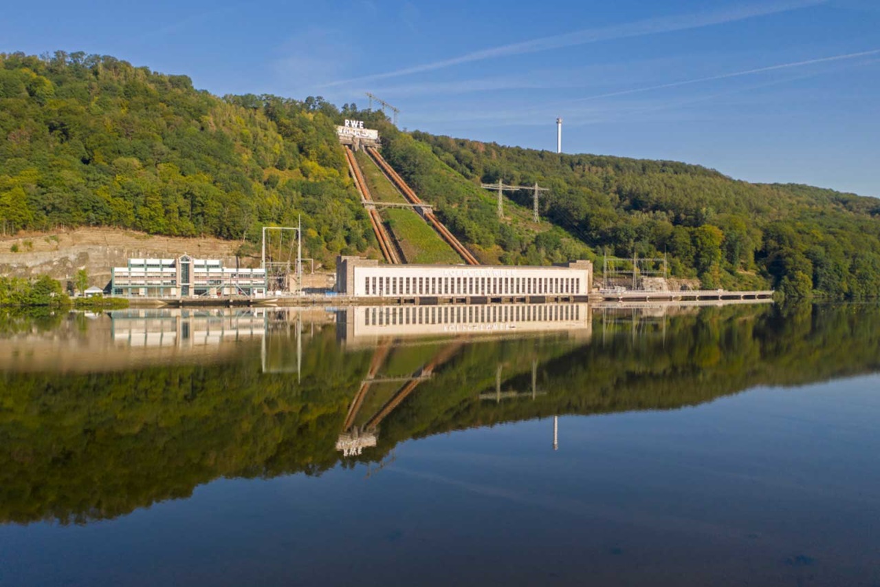 A hydroelectric power station by a calm lake, surrounded by wooded hills, reflecting in the water.